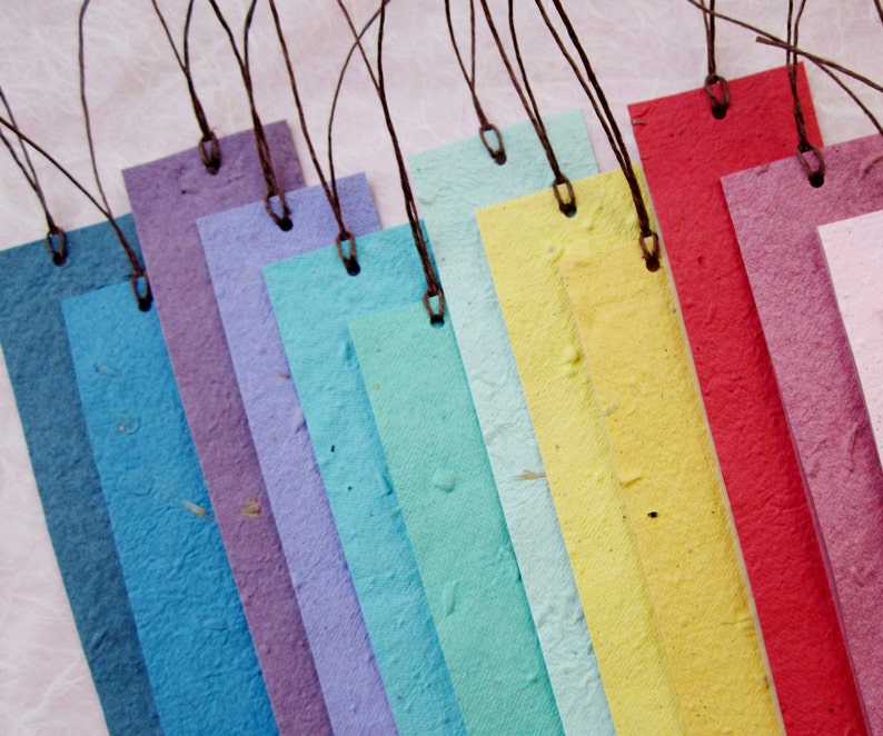 May include: A set of ten colorful bookmarks made from recycled paper with brown string attached. The bookmarks are arranged in a rainbow pattern, starting with white and ending with red.