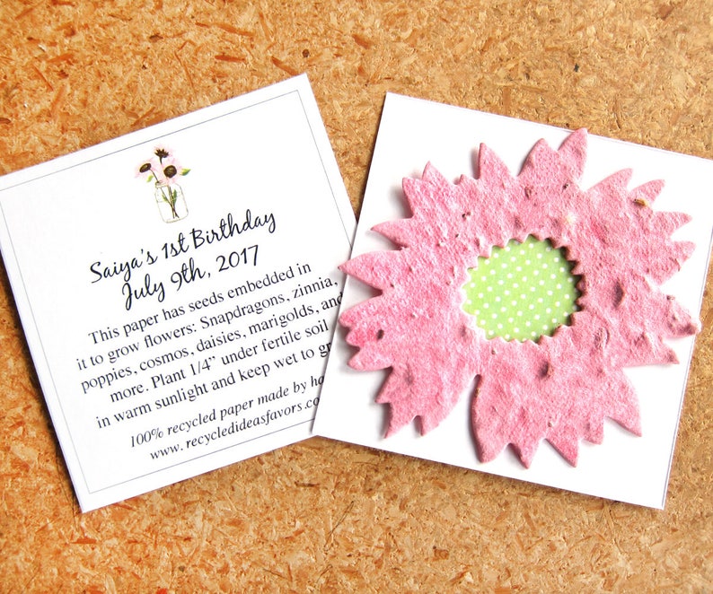 May include: A pink paper flower with a green polka dot center. The flower is a seed paper, which can be planted to grow flowers. The card next to the flower has text that reads "Saiya's 1st Birthday July 9th, 2017. This paper has seeds embedded in it to grow flowers: Snapdragons, zinnia, poppies, cosmos, daisies, marigolds, and more. Plant 1/4" under fertile soil in warm sunlight and keep wet to grow. 100% recycled paper made by hand. www.recycledideasfavors.com"