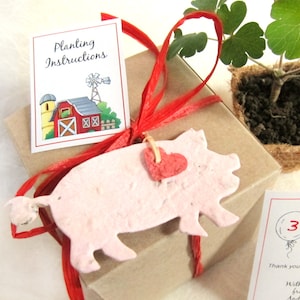 Puede incluir: Una caja de regalo marr&oacute;n con una cinta roja y un cerdo de papel rosa adjunto. El cerdo tiene un coraz&oacute;n rojo en un lado. La caja tiene una etiqueta que dice "Instrucciones de plantaci&oacute;n" con una ilustraci&oacute;n de dibujos animados de un granero rojo. La caja tambi&eacute;n tiene una etiqueta que dice "3" con un globo y el texto "&iexcl;Gracias por celebrar con nosotros!"