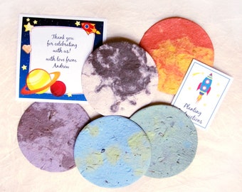 May include: Five round, colorful paper discs with a space theme. The discs are made of plantable paper and have different colors and patterns. There is a thank you card with a rocket and planets, and a card with planting instructions and a rocket.