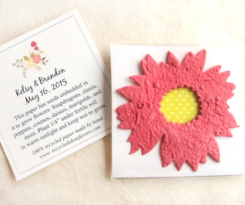 May include: A pink paper flower with a yellow center and white polka dots. The flower is packaged in a clear plastic bag with a white card that reads "Kelsy & Brandon May 16, 2015" and includes information about the flower seeds embedded in the paper.