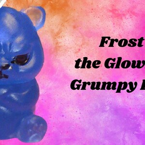 May include: A translucent blue bear figurine with a grumpy facial expression, wearing a white hat. The text "Frost the Glowing Grumpy Bear" is printed to the right, with a small "Evil Kitties" logo featuring a cat icon at the bottom right corner.