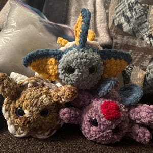 May include: Three handmade crochet plush toys in shades of brown, gray, and purple. The plushies have large, round eyes and various colored accents. The gray plush has yellow and blue accents, the purple has a red heart, and the brown has white accents.