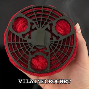 Op de afbeelding: Een rond, rood gehaakt item met een zwart, webachtig ontwerp aan de bovenkant, met kruisbeenderen. De tekst "VILAINE CROCHET" is zichtbaar aan de onderkant. Het item lijkt handgemaakt.