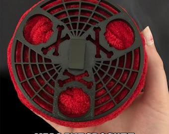 May include: A round, red crochet item with a black, web-like design on top, featuring crossbones. The text "VILAINE CROCHET" is visible at the bottom. The item appears to be handmade.