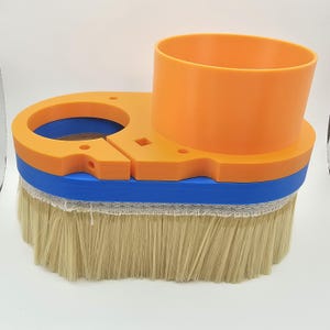 May include: A close-up of a router brush attachment with a blue and orange base and a beige brush. The brush is designed for use with a router to smooth and finish wood.