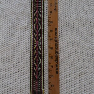May include: A woven textile strip with a geometric pattern in black, burgundy, yellow, and white. The strip is approximately 13 inches long. The design includes diamond shapes and fringe at both ends. A wooden ruler is next to the strip.