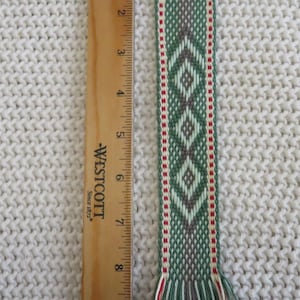 May include: A woven bookmark featuring a geometric diamond pattern in shades of green, grey, and cream with red accents, finished with fringed ends. A wooden Westcott ruler is placed alongside, showing the bookmark is 9 inches long.