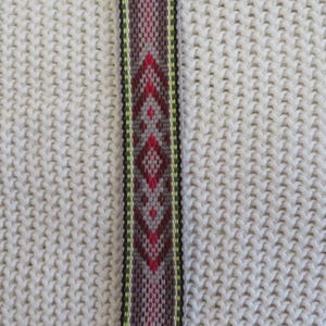 May include: A woven bookmark featuring a geometric pattern in shades of red, gray, and lime green. The design includes diamond shapes centered along the length, finished with fringed ends made of loose, multi-colored yarn strands.