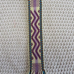 May include: A woven bookmark featuring a cream-colored base with a central purple zigzag pattern. The design includes vertical stripes of bright blue and black, finished with loose, multi-colored fringe at both the top and bottom ends.