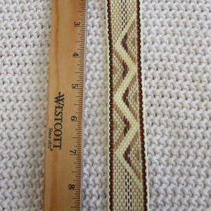 May include: A woven bookmark with a cream, tan, and brown geometric chevron pattern, featuring fringed ends and thin red and black border stripes. A wooden Westcott ruler is placed alongside, showing the bookmark is 10 inches long.