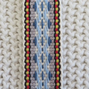 May include: A woven fabric strap featuring a central geometric pattern in light blue, dark blue, and gray, flanked by vertical stripes of red, yellow, and black, set against a white knitted background with a visible textured stitch pattern.