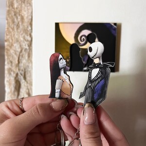 May include: A keychain featuring Jack Skellington and Sally from the Nightmare Before Christmas. The characters are depicted in a cartoon style and are holding hands. The keychain is attached to a silver chain and a set of keys.
