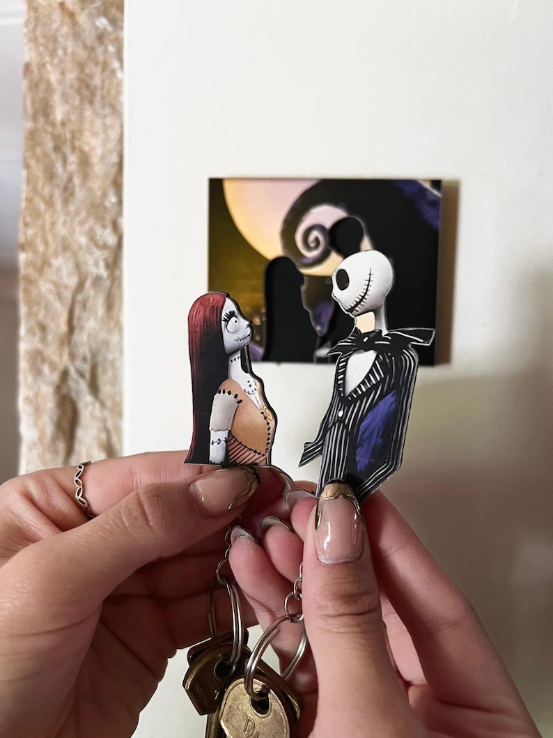 May include: A keychain featuring Jack Skellington and Sally from the Nightmare Before Christmas. The characters are depicted in a cartoon style and are holding hands. The keychain is attached to a silver chain and a set of keys.