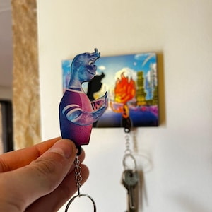 May include: A blue and purple character keyring with a key ring attached. The character is holding a small object in its hand. The keyring is hanging from a key hook with other keys.