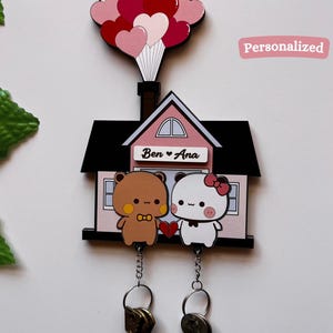 May include: A personalized key holder featuring a house design with two cartoon bears, one brown and one white, holding a heart. The house has a black roof and a pink facade, with the names "Ben & Ana" above the bears. A cluster of heart-shaped balloons tops the house.