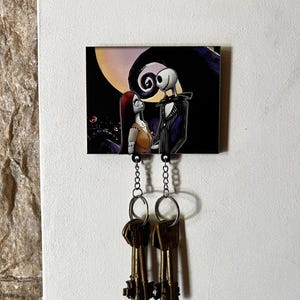 May include: A key holder featuring a black and white illustration of Jack Skellington and Sally from the Nightmare Before Christmas movie. The illustration is on a black background with a white moon and a swirl design. Two keys are hanging from the key holder.