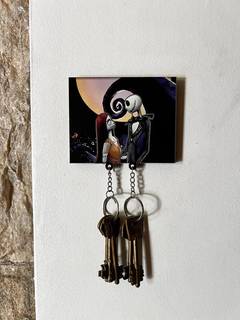 May include: A key holder featuring a black and white illustration of Jack Skellington and Sally from the Nightmare Before Christmas movie. The illustration is on a black background with a white moon and a swirl design. Two keys are hanging from the key holder.