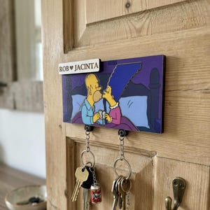May include: A rectangular wall-mounted key holder featuring a printed illustration of Homer and Marge Simpson kissing in bed. A small white plaque attached to the top displays the text ROB ♥ JACINTA. Two metal hooks hold silver key rings with keys.