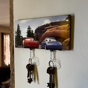 Może przedstawiać: Klucznik z nadrukowanym obrazkiem dwóch kreskówek samochodów, jednego czerwonego i jednego niebieskiego, jadących po górskiej drodze. Klucznik ma dwa haczyki z wiszącymi kluczami.