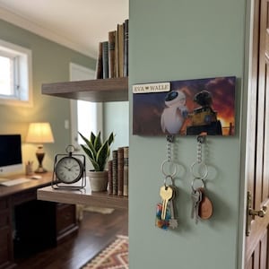 Puede incluir: Un soporte de pared rectangular para llaves con una imagen impresa de los personajes EVE y WALL-E, y una pequeña placa superior con la inscripción EVA ♥ WALLE. Incluye dos ganchos metálicos plateados, cada uno con un llavero de metal.