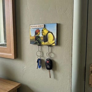 May include: A rectangular wall-mounted key holder featuring a printed image of Shrek and Fiona with the text FIONA HEART SHREK. The wooden plaque has two metal hooks at the bottom, each holding a silver keyring with attached keys and a car fob.