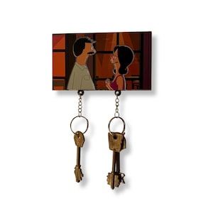 May include: A wooden key holder with a cartoon image of two people looking at each other. The key holder has two hooks with keys hanging from them.