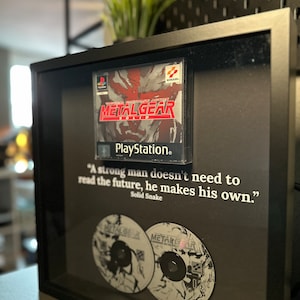 Könnte beinhalten: Eine gerahmte Präsentation mit einer PlayStation-Spielkassette für Metal Gear Solid, zwei Spielscheiben und einem Zitat aus dem Spiel: "Ein starker Mann muss die Zukunft nicht lesen, er macht sie selbst." - Solid Snake
