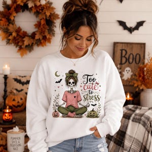 Puede incluir: Una sudadera blanca de manga larga con un gráfico de un esqueleto en posición de yoga. El esqueleto viste una camiseta rosa, leggings verdes y una diadema verde. El texto dice Too cute to stress, rodeado de calabazas, murciélagos y corazones.