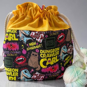 May include: A drawstring project bag with a bright yellow top and a black base featuring a colorful graphic print. The print includes text reading Dungeon Crawler Carl, New Achievement, and God Damn Donut, alongside illustrations of a cat and dinosaurs.