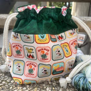 May include: A drawstring project bag featuring a dark green fabric top and a white base printed with a repeating pattern of rectangular patches depicting Mickey and Minnie Mouse, flowers, and mountains. The bag has thick white rope drawstrings.