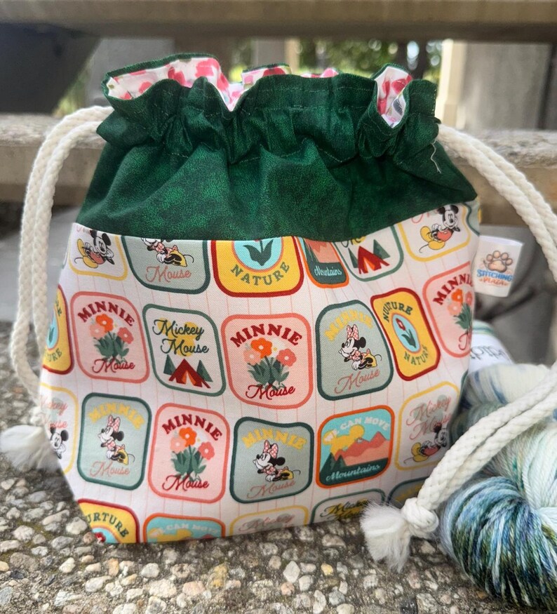 May include: A drawstring project bag featuring a dark green fabric top and a white base printed with a repeating pattern of rectangular patches depicting Mickey and Minnie Mouse, flowers, and mountains. The bag has thick white rope drawstrings.