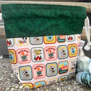 May include: A drawstring bag featuring a dark green top band and a white base patterned with colorful rectangular patches. Each patch displays illustrations of Mickey and Minnie Mouse, flowers, mountains, and tents, with text including "Minnie Mouse," "Nature," and "Mountains."