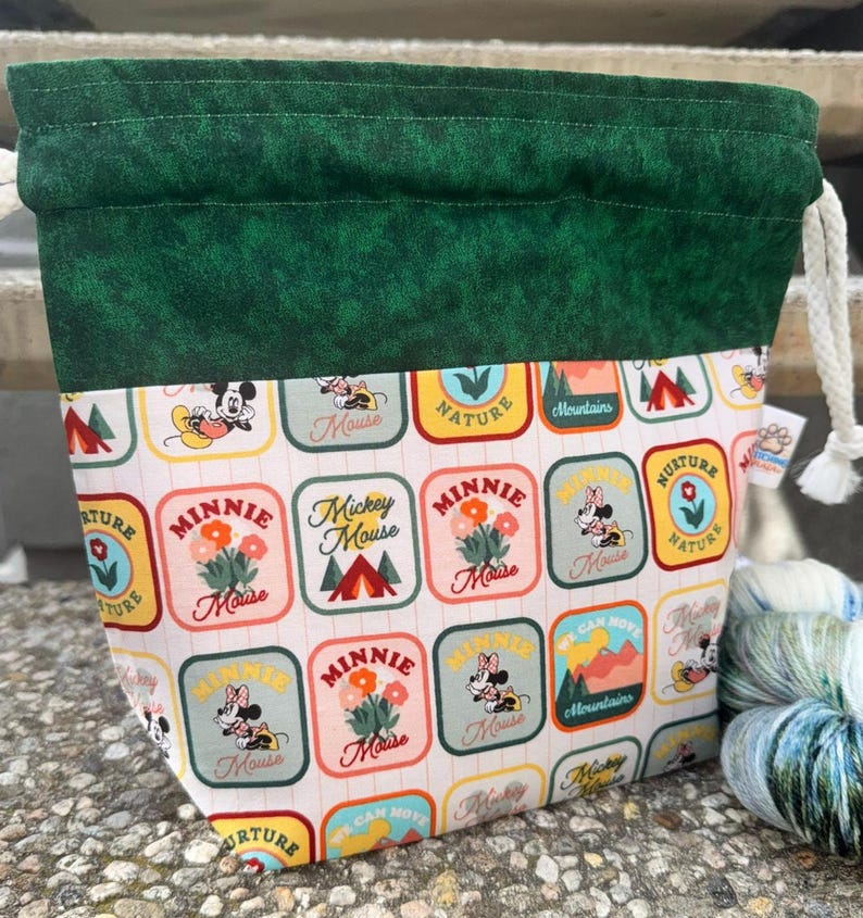 May include: A drawstring bag featuring a dark green top band and a white base patterned with colorful rectangular patches. Each patch displays illustrations of Mickey and Minnie Mouse, flowers, mountains, and tents, with text including "Minnie Mouse," "Nature," and "Mountains."