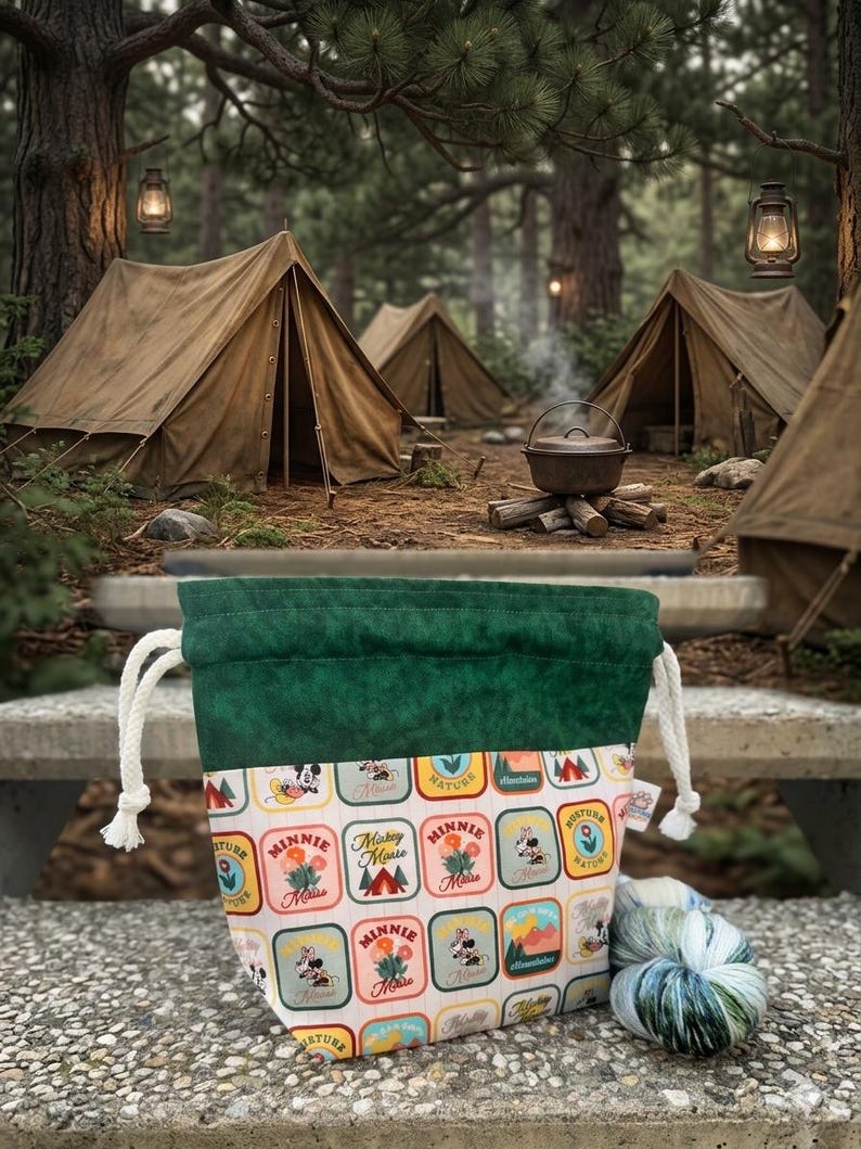 May include: A drawstring pouch featuring a dark green top band and a white lower section with a repeating grid pattern of colorful patches depicting Minnie Mouse, Mickey Mouse, flowers, and mountains, accompanied by a skein of variegated yarn.