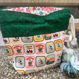 May include: A drawstring project bag with a dark green fabric top and a white base featuring a repeating pattern of colorful rectangular patches with Mickey and Minnie Mouse, nature, and mountain graphics. The interior lining has a floral print.