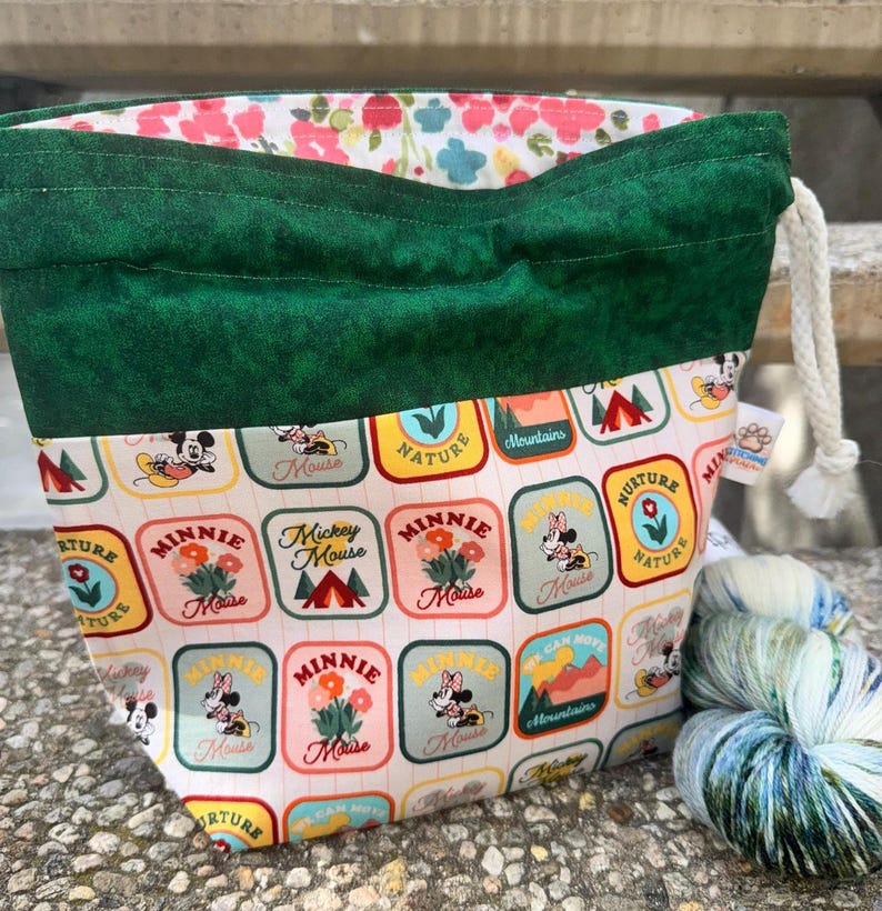 May include: A drawstring project bag with a dark green fabric top and a white base featuring a repeating pattern of colorful rectangular patches with Mickey and Minnie Mouse, nature, and mountain graphics. The interior lining has a floral print.