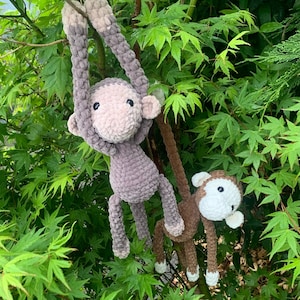 Pode incluir: Dois brinquedos de macaco de crochê pendurados em um galho de árvore. O macaco maior é cinza e o macaco menor é marrom com detalhes brancos.