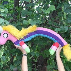 May include: A plush rainbow unicorn toy with a white horn, yellow mane and tail, and a pink body. The toy has a rainbow-colored body and is held up against a background of green leaves.