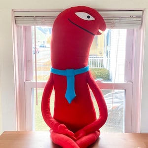 Puede incluir: Una figura de peluche roja grande con un cuerpo largo, brazos y piernas, sentada con las piernas cruzadas. Tiene una corbata azul y un ojo blanco. La figura est&aacute; sentada sobre una superficie de madera frente a una ventana.