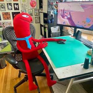 Puede incluir: Un gran peluche rojo con una corbata azul est&aacute; sentado en una silla de oficina negra en un escritorio. El juguete tiene brazos y piernas largos, y un solo ojo. Un monitor de computadora y &uacute;tiles de arte son visibles en el escritorio.