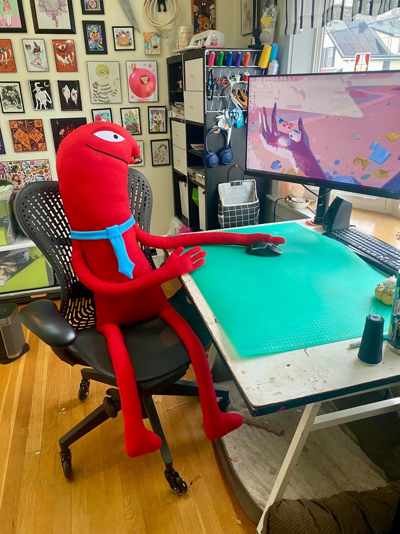 Puede incluir: Un gran peluche rojo con una corbata azul est&aacute; sentado en una silla de oficina negra en un escritorio. El juguete tiene brazos y piernas largos, y un solo ojo. Un monitor de computadora y &uacute;tiles de arte son visibles en el escritorio.
