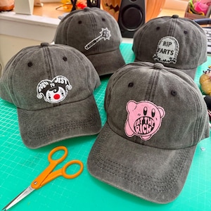May include: Four charcoal gray baseball caps with embroidered designs. One features a jester face with the text BORN TO JEST, one a spiked mace, one a tombstone with RIP FARTS, and one a pink character with the text EAT THE RICH. Orange scissors are visible.
