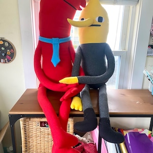 Puede incluir: Dos figuras de peluche sentadas sobre una mesa de madera. Una es roja con una corbata azul, la otra es amarilla y gris. La figura roja tiene un cuerpo largo y curvado y un solo ojo. La figura amarilla tiene una nariz larga y una cara cuadrada.