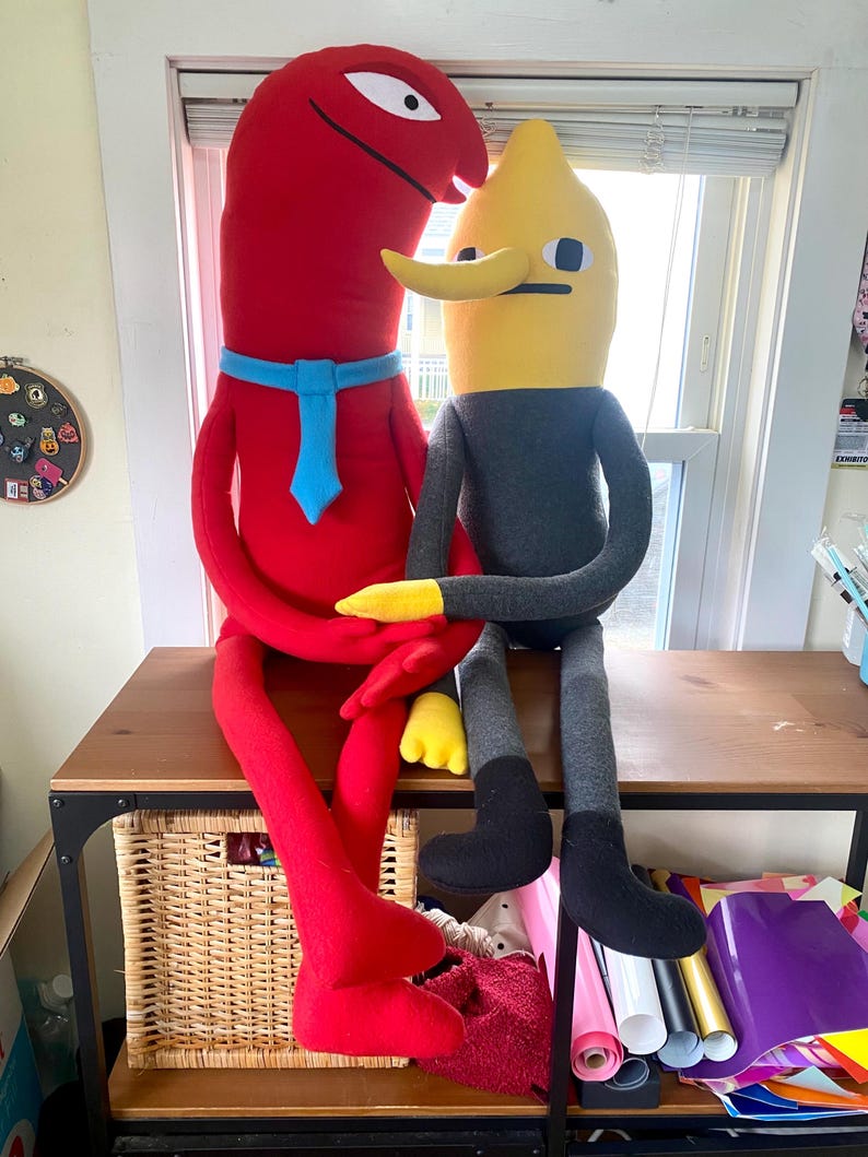 Puede incluir: Dos figuras de peluche sentadas sobre una mesa de madera. Una es roja con una corbata azul, la otra es amarilla y gris. La figura roja tiene un cuerpo largo y curvado y un solo ojo. La figura amarilla tiene una nariz larga y una cara cuadrada.