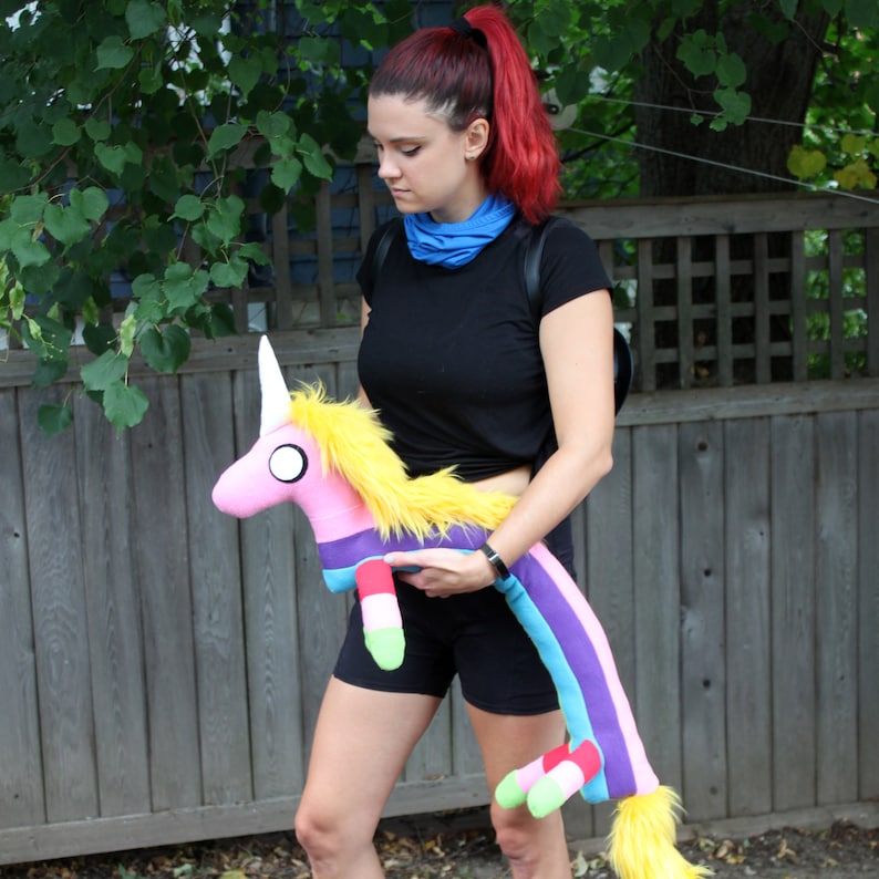 May include: A plush unicorn toy with a pink body, yellow mane, and a long, striped tail in shades of purple, blue, and red. The unicorn has a white horn and large, round eyes. The toy is being held in front of a wooden fence.