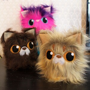 May include: Three plush toy cats with large eyes. The cat on the left is brown, the cat in the middle is pink with purple fur, and the cat on the right is brown and tan.