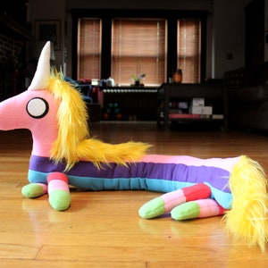 May include: A plush, elongated unicorn toy with a pink body, yellow mane and tail, and a white horn. The unicorn features a rainbow-striped body and green and red feet. The toy is lying on a wooden floor.