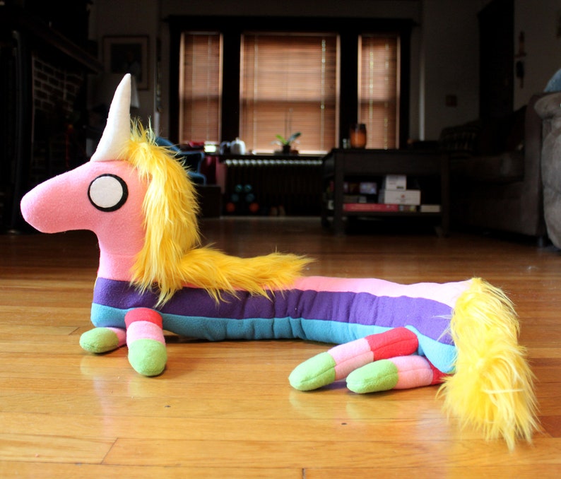 May include: A plush, elongated unicorn toy with a pink body, yellow mane and tail, and a white horn. The unicorn features a rainbow-striped body and green and red feet. The toy is lying on a wooden floor.