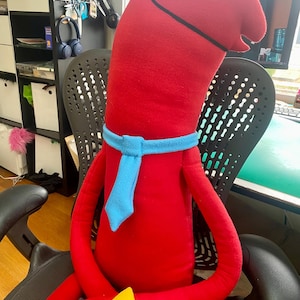 Puede incluir: Un personaje de peluche rojo con una bufanda azul y un tri&aacute;ngulo amarillo. El personaje tiene un cuerpo largo y delgado, una cabeza grande con un ojo blanco y est&aacute; sentado en una silla negra. Los brazos del personaje descansan sobre su regazo.